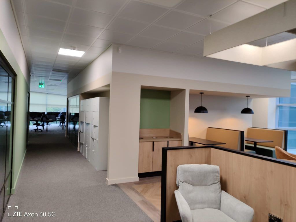 Modern office hallway featuring a beige partition, white storage lockers, light wood cabinetry, and a cozy lounge area.