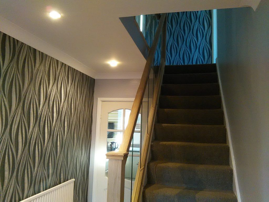 A hallway with patterned wallpaper on the left, a white radiator, a staircase with dark carpet, and a glass-paneled door.