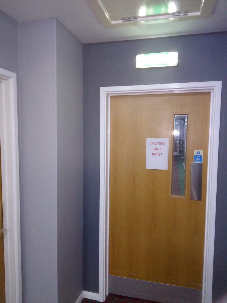 A light-wood door with a narrow window and a white sign sits in a grey hallway under an emergency exit light.