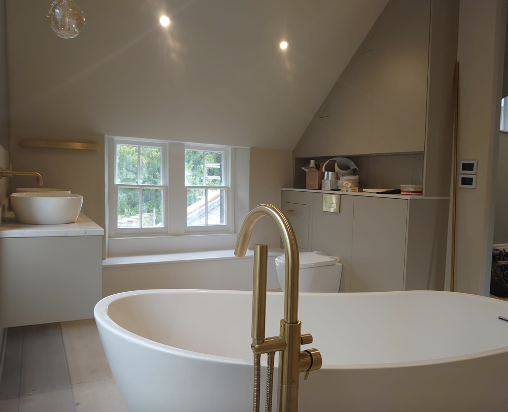 A modern, neutral-toned bathroom featuring a white freestanding bathtub with gold fixtures and a built-in window seat.