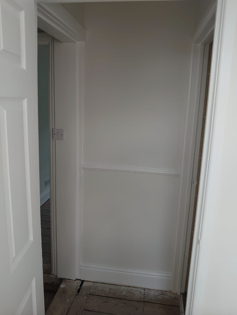 An indoor view of a pale gray hallway with white trim, a chair rail, and an open doorway leading to another room.