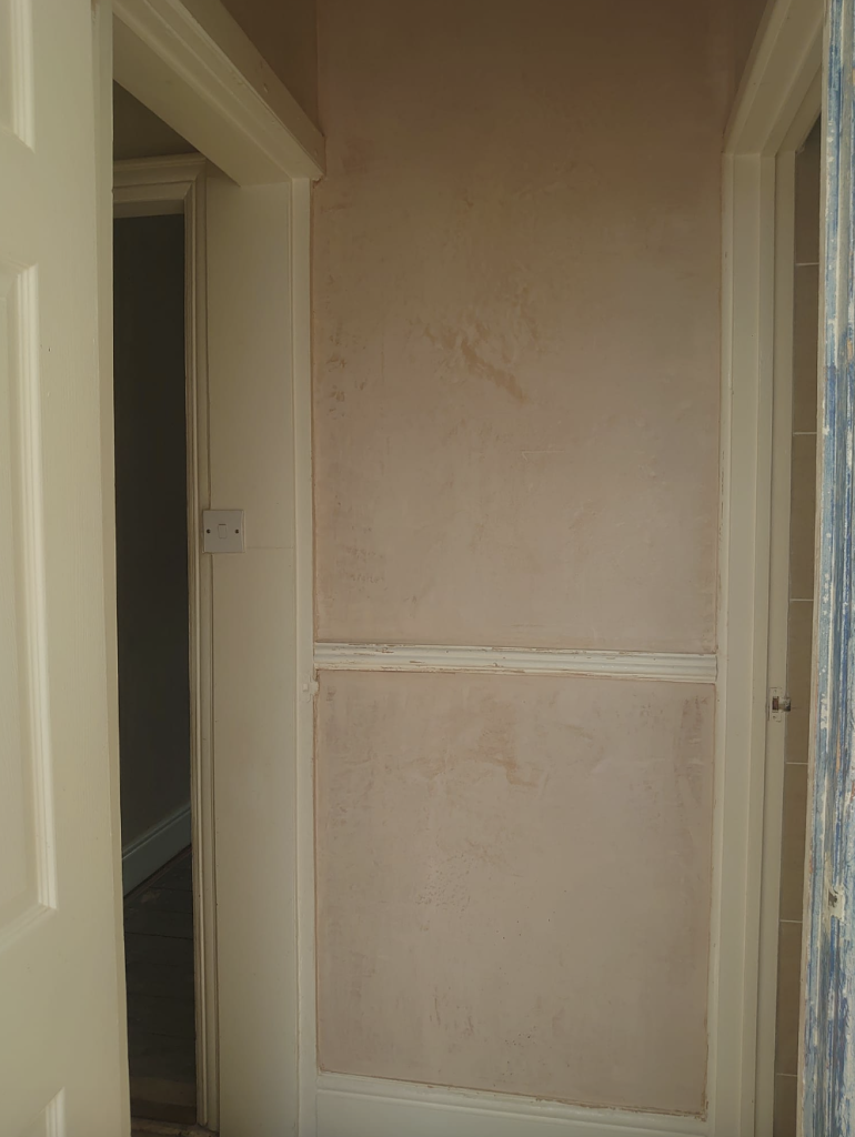 A view of a hallway wall featuring a rectangular section of newly plastered, light brown wallboard set between door frames.