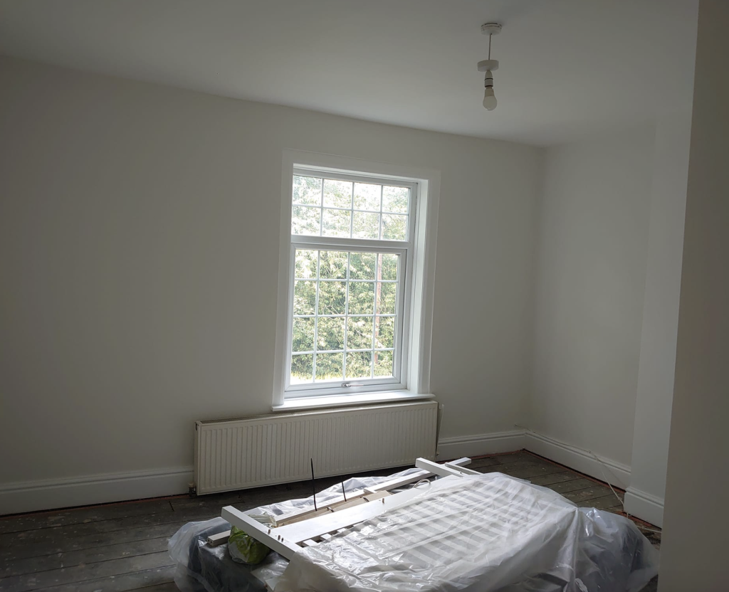 An empty room with white walls, a large window, a radiator, and a covered item in the foreground.