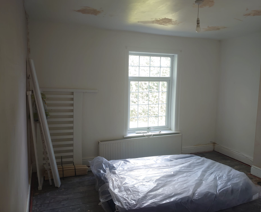 A room under renovation with white walls, a plastic-covered floor, a radiator, and a window with water-stained ceiling.