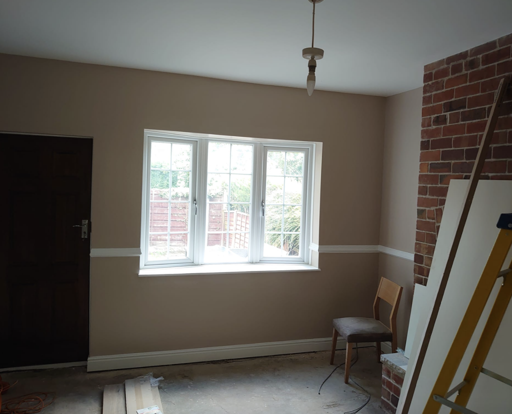An unfinished room with beige walls, a window, a chair, and an exposed brick fireplace chimney breast with a ladder.