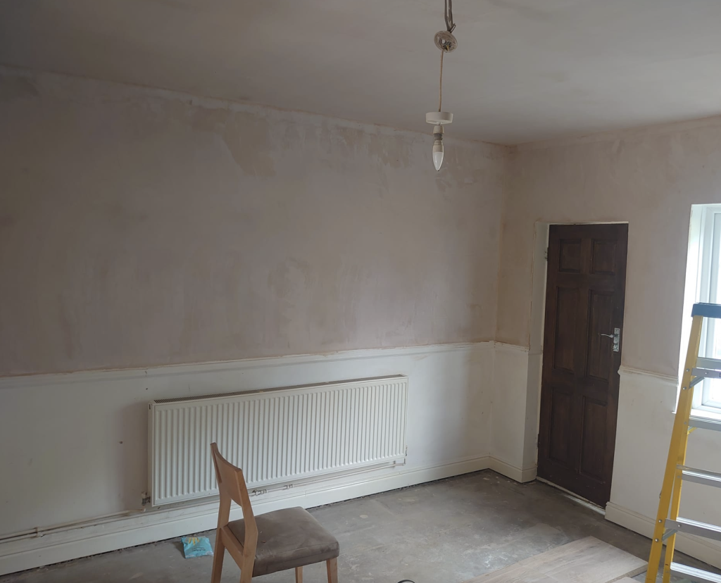 A room under renovation with partially painted walls, a radiator, a wooden chair, a dark door, and a yellow stepladder.