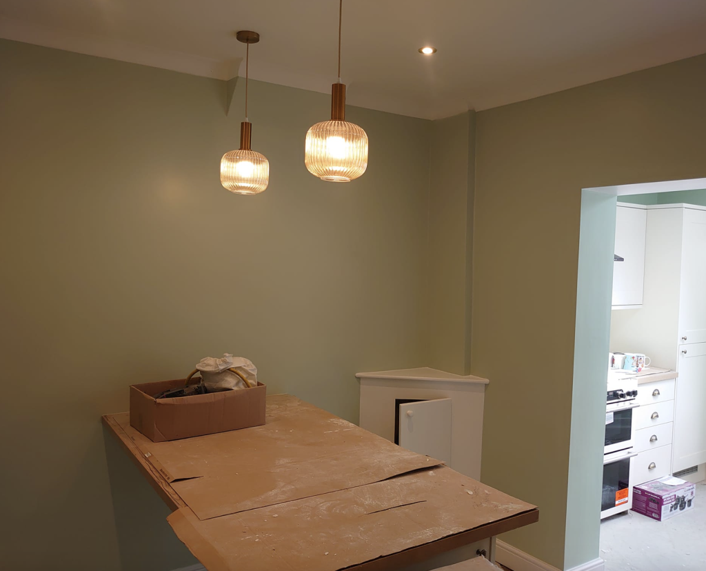 A light green room with two hanging pendant lights over a wooden table, featuring a partial view of a kitchen.