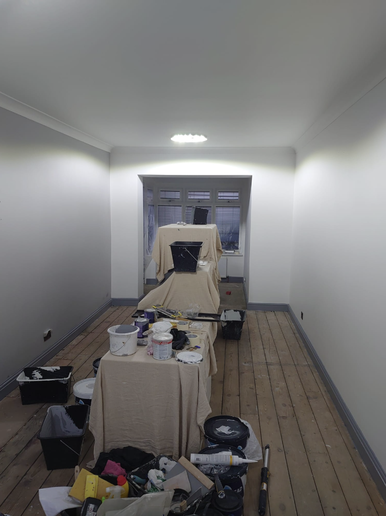 A long, narrow room under renovation with freshly painted white walls, bare wooden floorboards, and covered furniture.