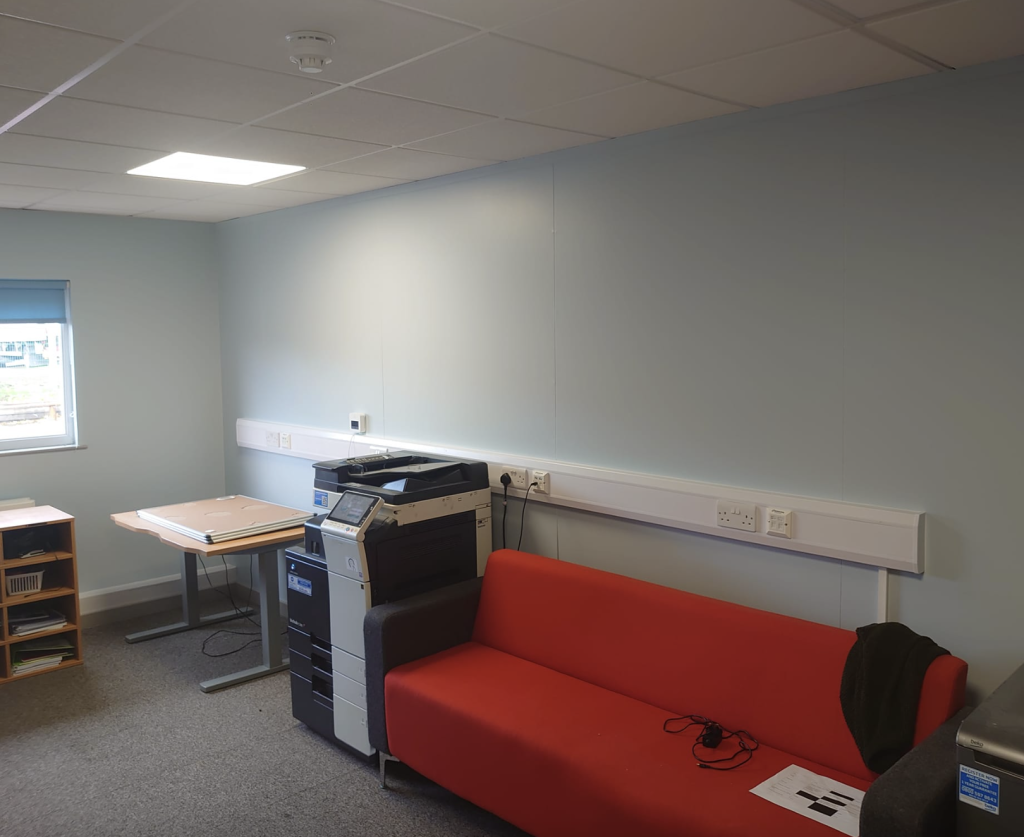 A small office space with a red sofa, a printer, a desk, a shelving unit, and a window on a light blue wall.