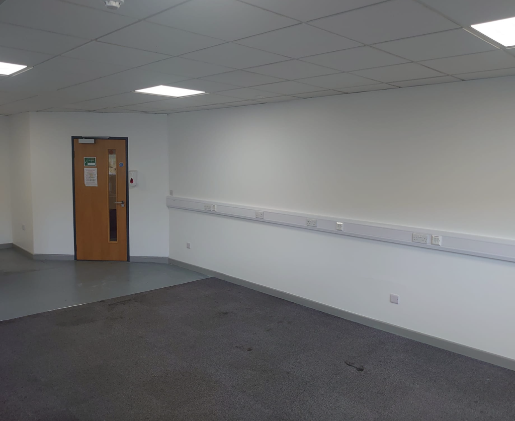 A wide, empty office space with gray carpet, white walls, overhead lighting, and a wooden door on the left.