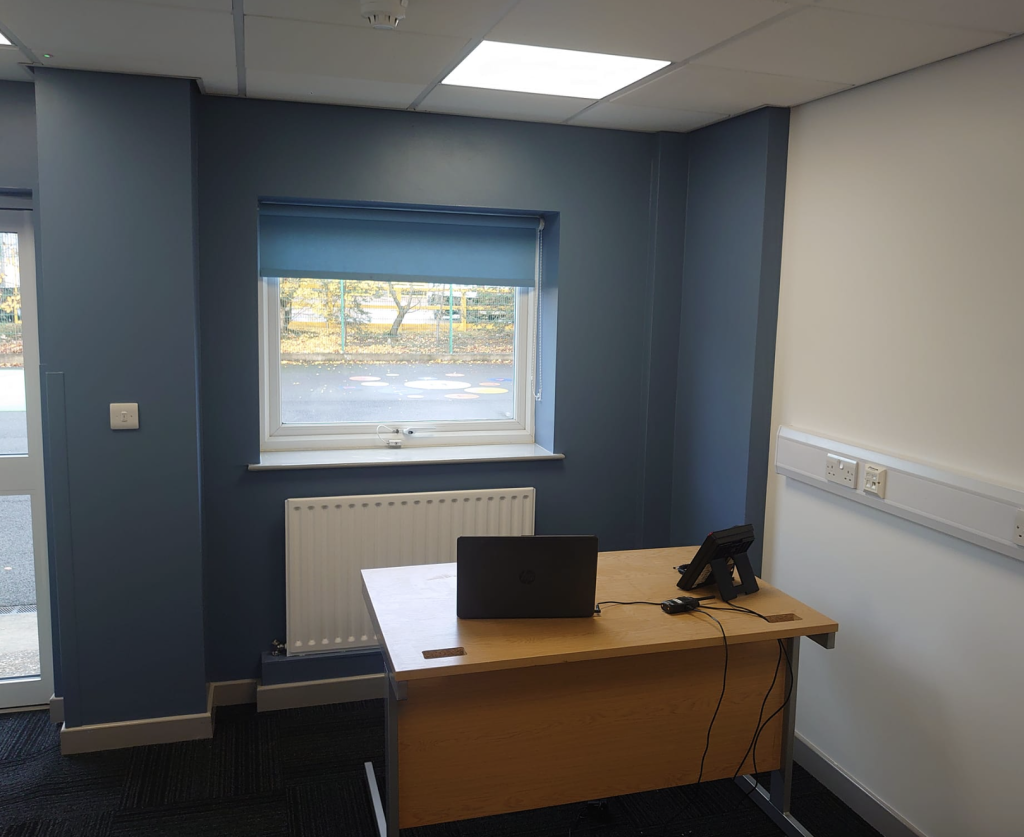 A small office with a wooden desk, a laptop, and a telephone, set against blue and white walls with a window.