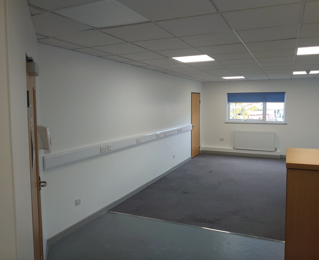 An empty office space with white walls, grey carpeting, a large window, a radiator, and drop ceiling lights.