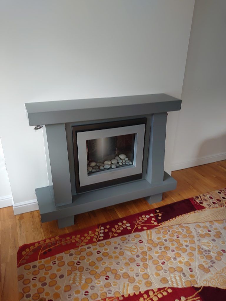 A grey, modern freestanding fireplace with a square insert filled with white decorative stones, set on a hardwood floor.