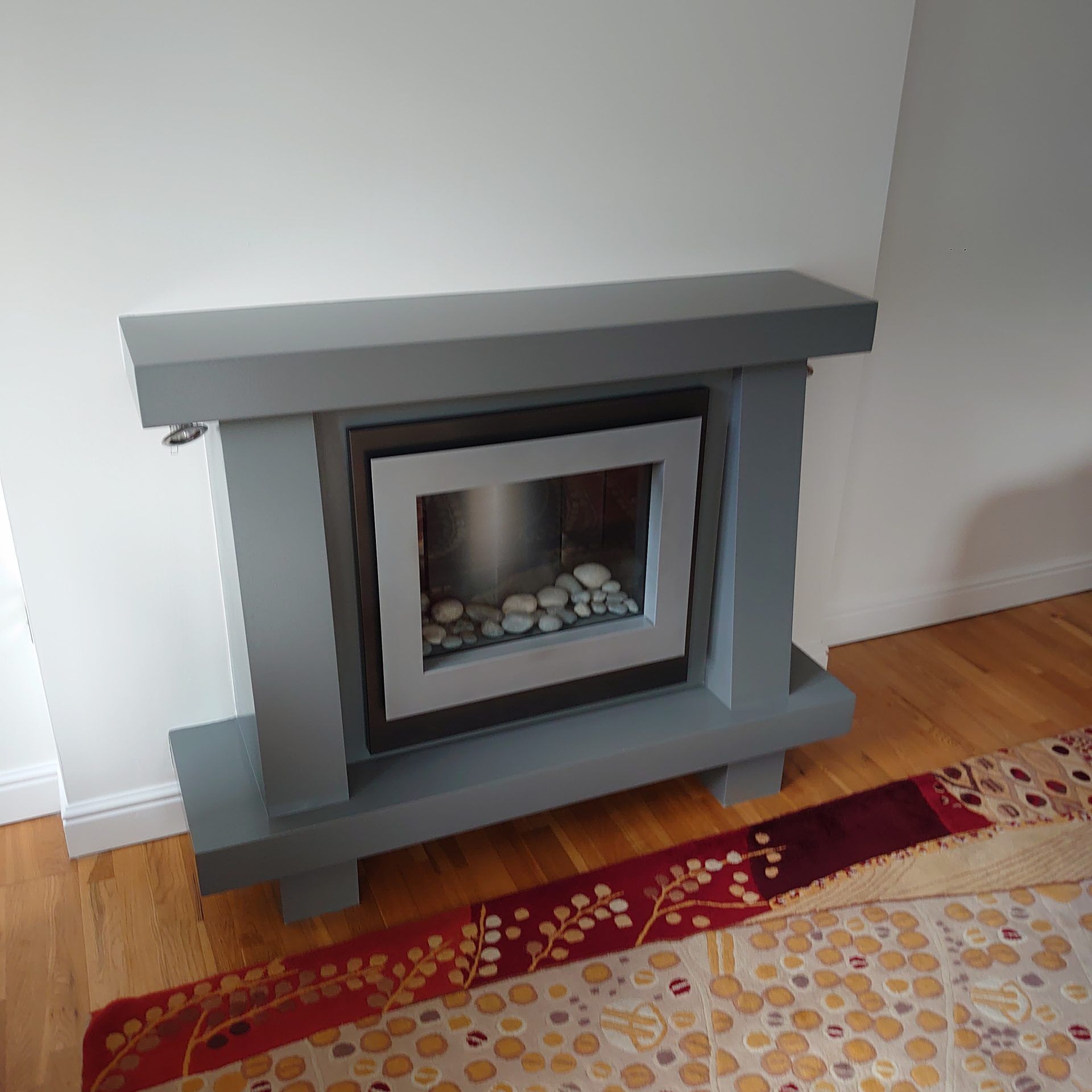 A gray, modern, freestanding electric fireplace with a decorative light gray frame and white pebbles, on a wooden floor.