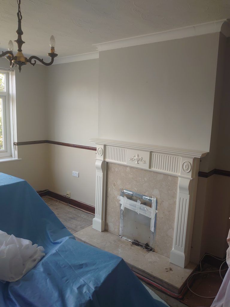 A room featuring a white ornamental fireplace surround, neutral walls with a dark trim, and furniture covered in blue tarps.