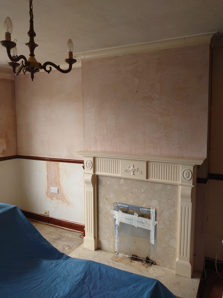 A fireplace with a light-colored mantel in a room with partially replastered pinkish walls and a hanging chandelier.