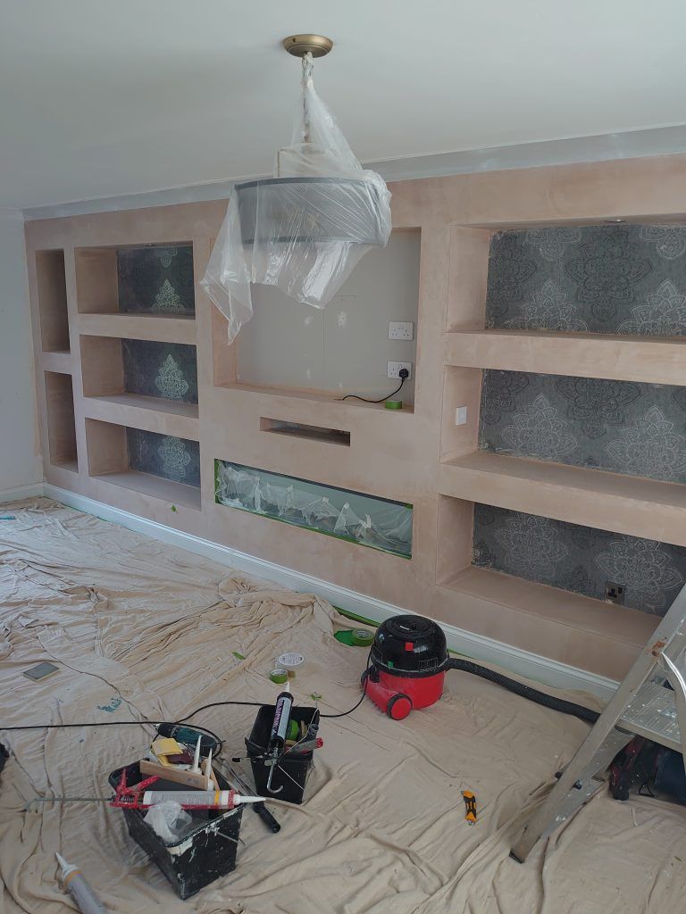 A newly plastered media wall with built-in shelves under construction, surrounded by tools and drop cloths in a room.