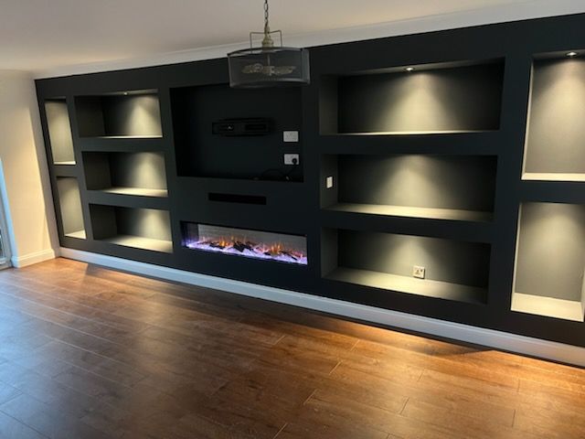 A matte black media wall featuring built-in recessed shelving, a central fireplace, and integrated spotlighting.