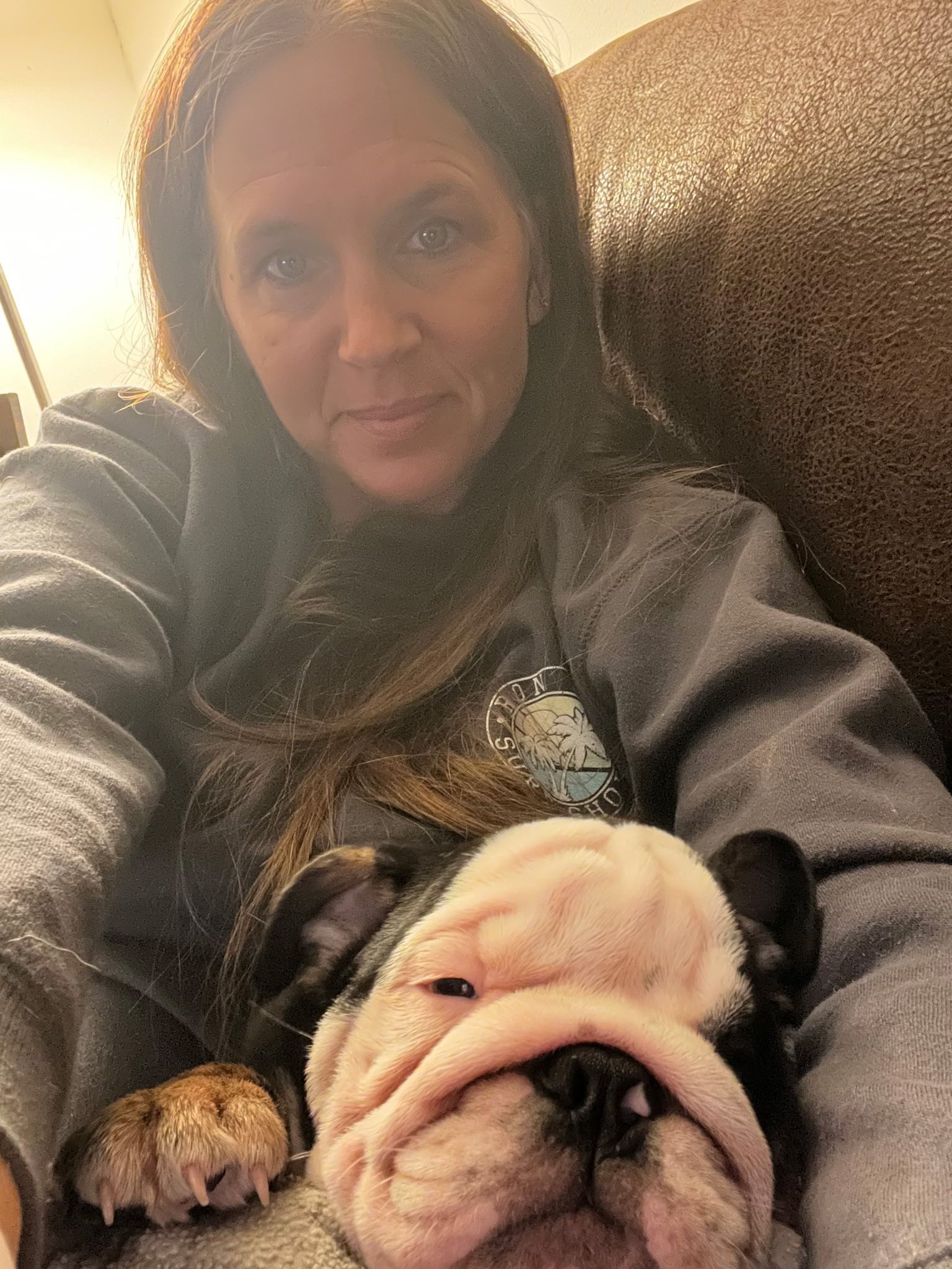 Woman with long brown hair and gray sweatshirt cuddles a sleeping bulldog on a couch.