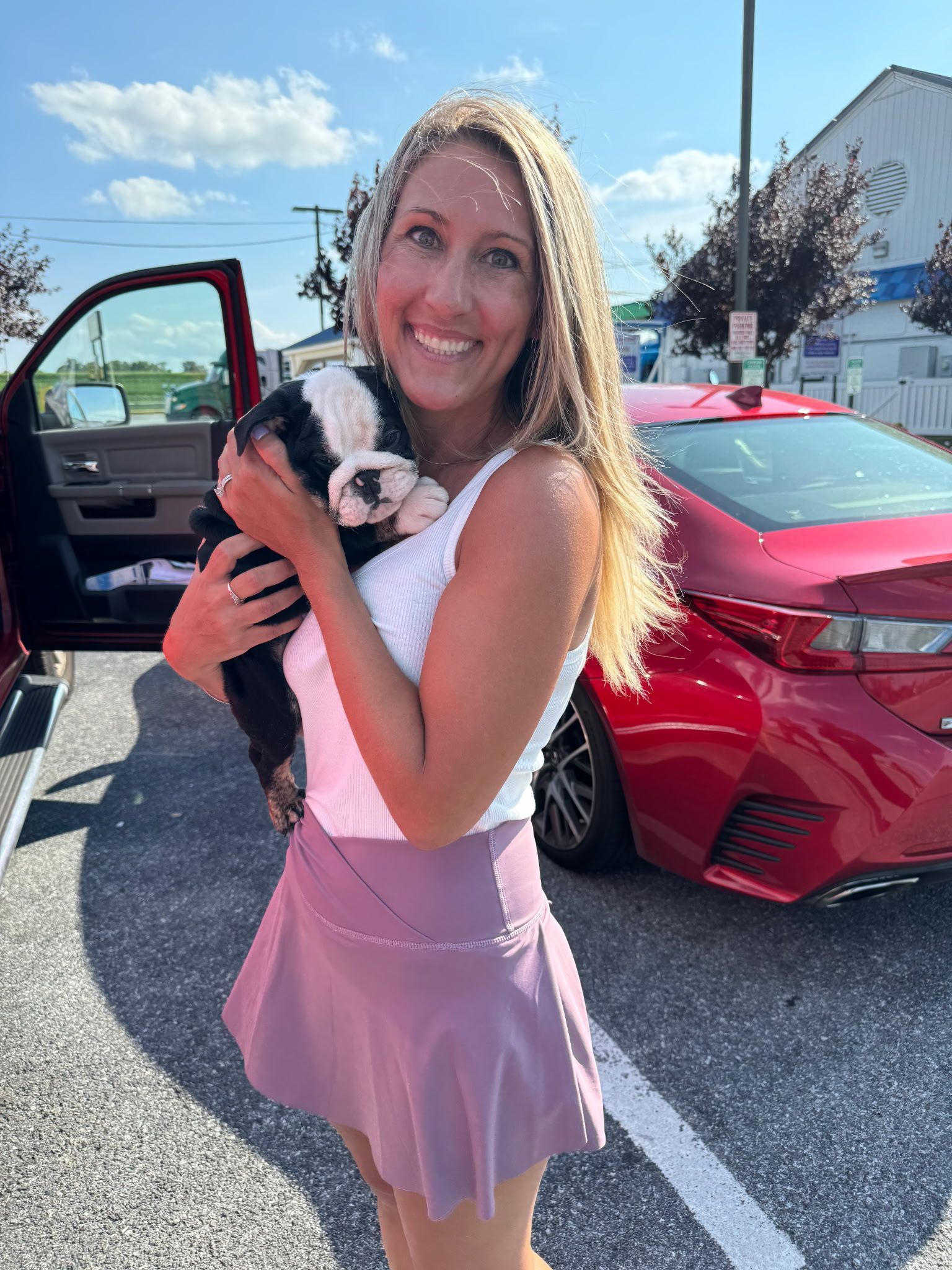 Woman with long blonde hair holds a black and white puppy while smiling outdoors, near a red car.