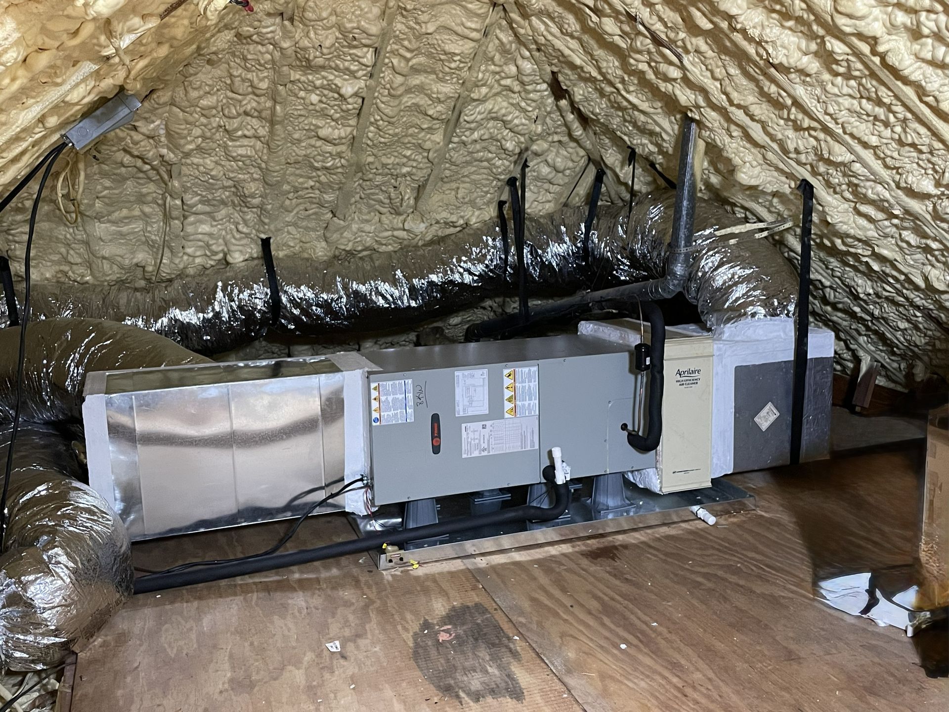 HVAC air handler unit installed in an attic with spray foam insulation on the roof rafters and exposed ductwork.