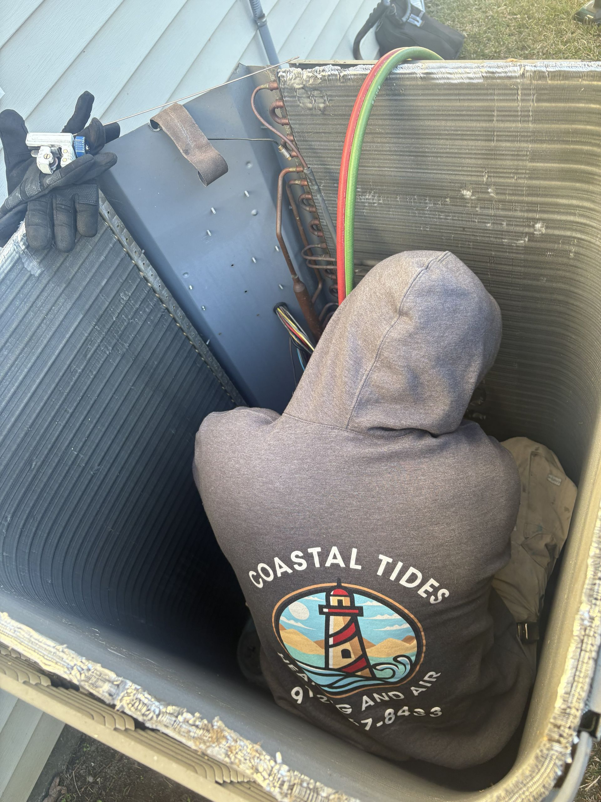 A technician in a hooded sweatshirt works inside an opened HVAC condenser unit, connecting gauge hoses to the coils.