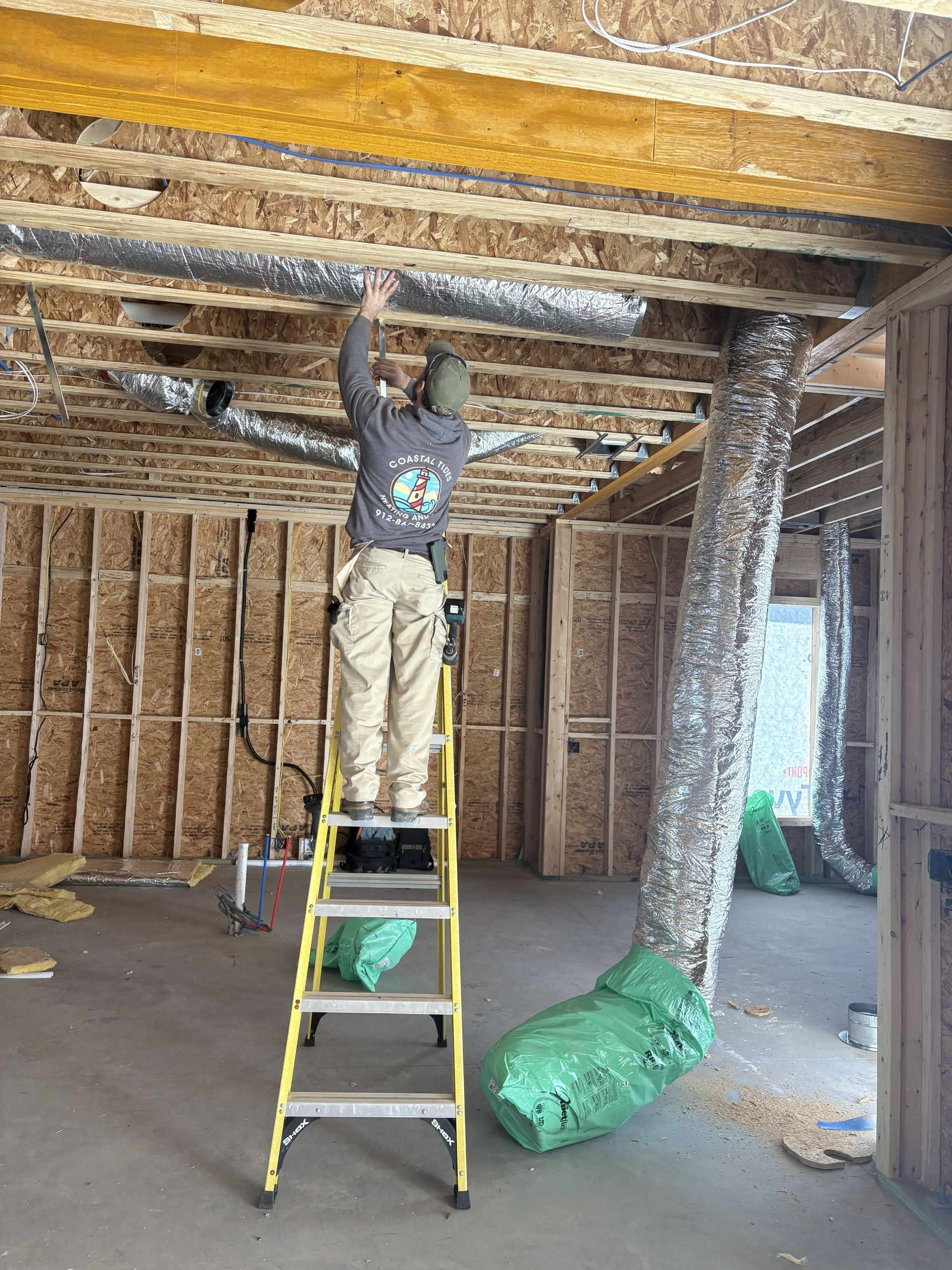 A person on a ladder installs silver flexible ductwork in the exposed wooden ceiling of an unfinished construction site.