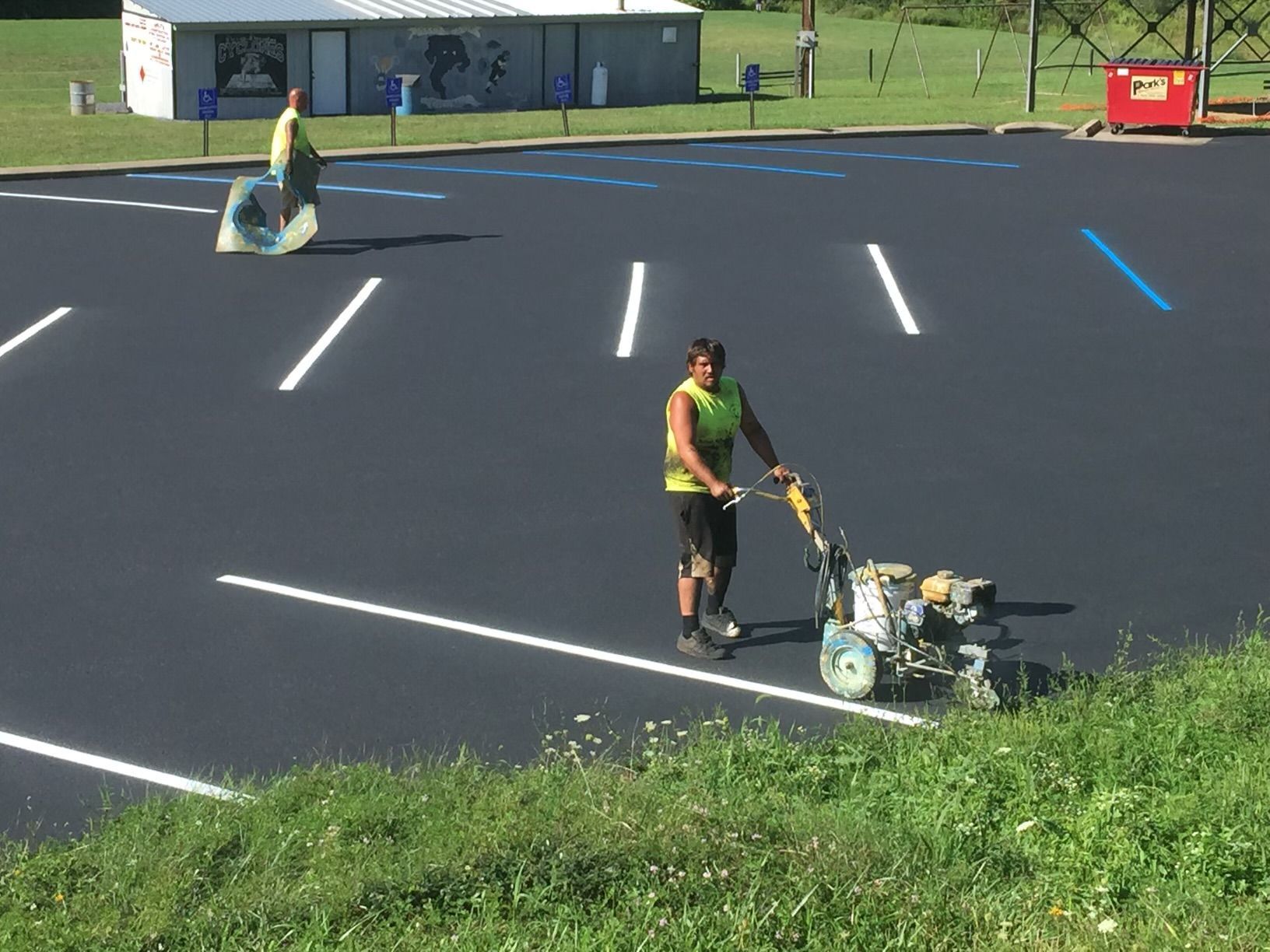 Workers painting parking lot lines with striping machine and collecting trash.