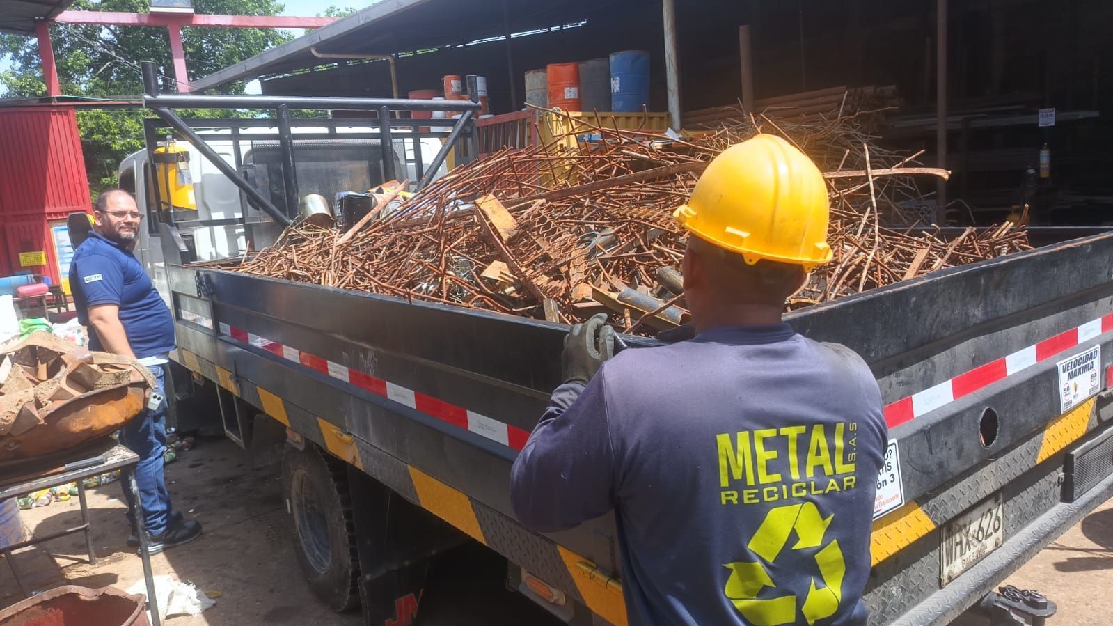 Dos hombres cargando la caja de un camión llena de chatarra; uno de ellos lleva un casco amarillo.