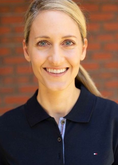 A woman wearing a black polo shirt is smiling in front of a brick wall.
