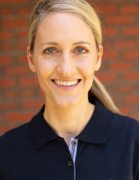 A woman wearing a black polo shirt is smiling in front of a brick wall.
