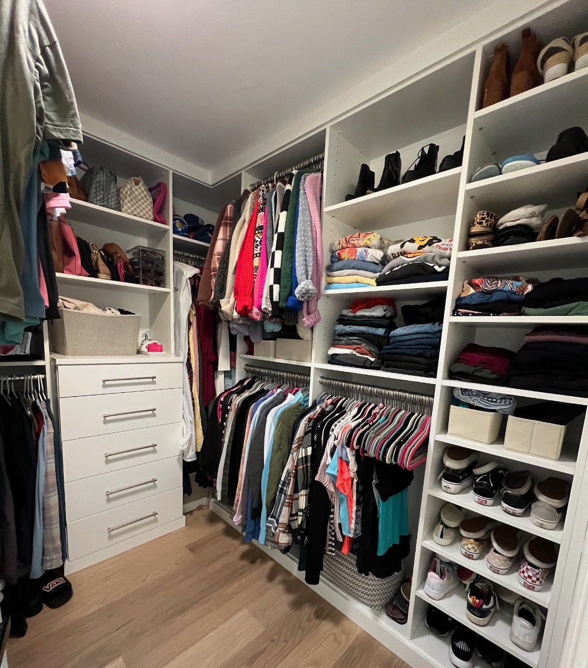 Walk-in closet with white shelves, hanging clothes, stacked clothes, and shoes.