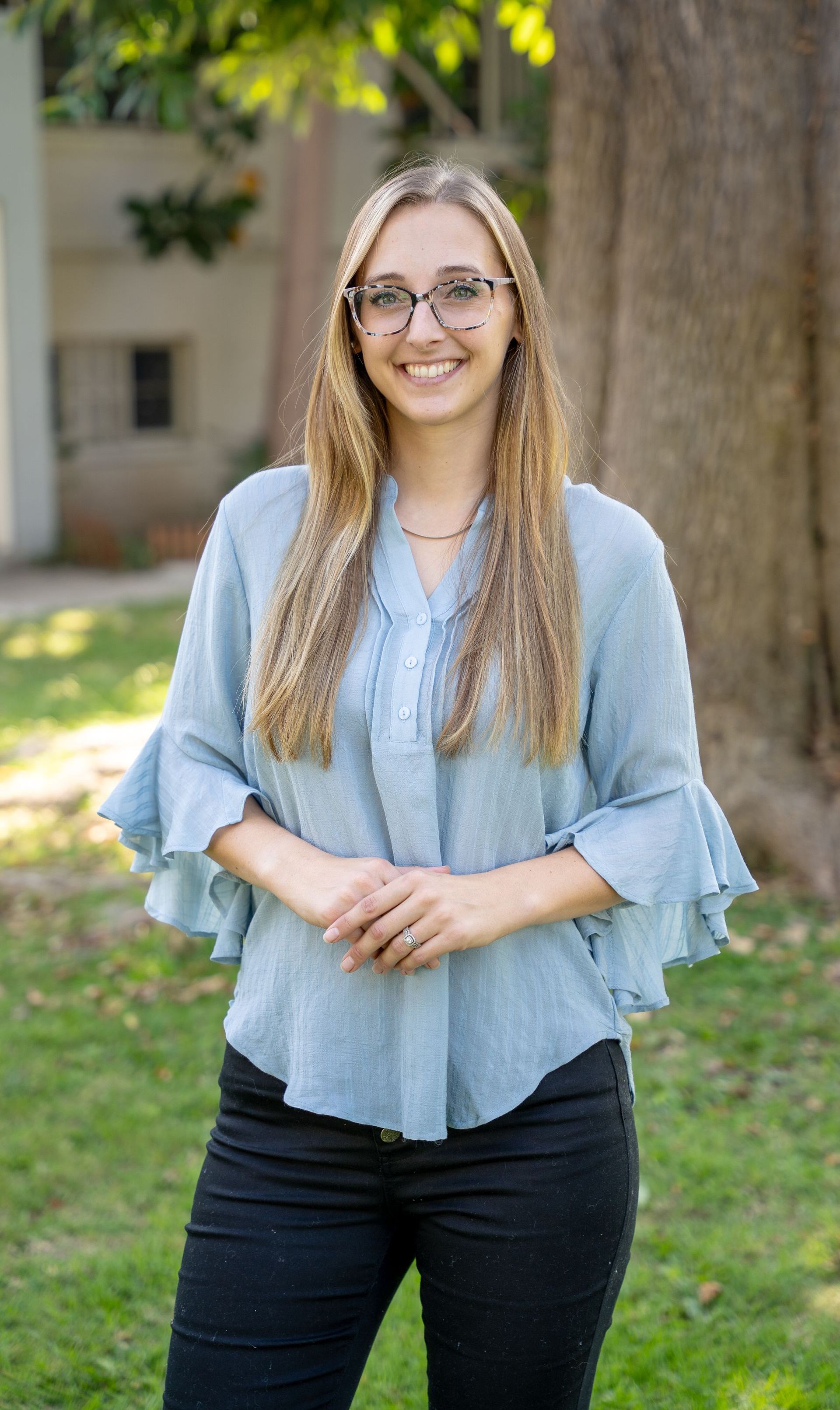 A woman wearing glasses and a blue shirt is standing in front of a tree.