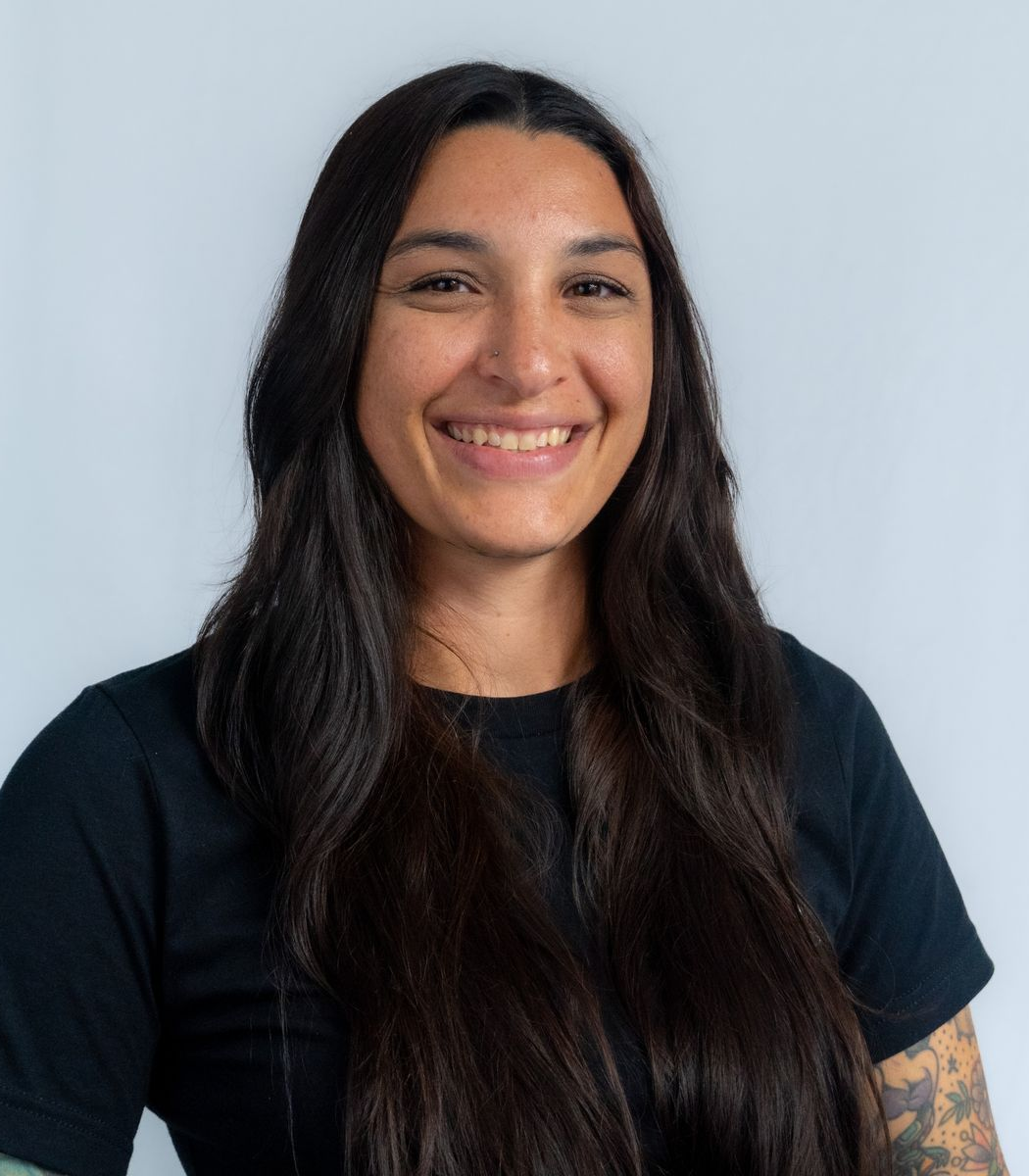 Smiling person with dark hair and a black beanie; outside with mountains in the background.