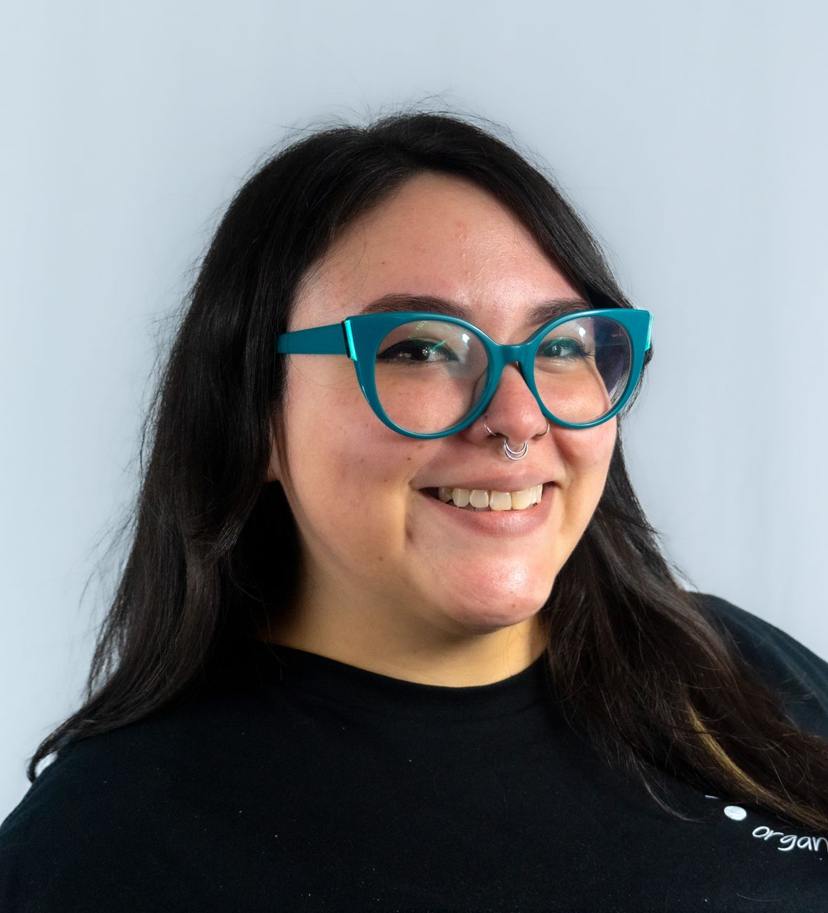 Woman with glasses, black hair with pink streaks, smiling in front of a white wall.