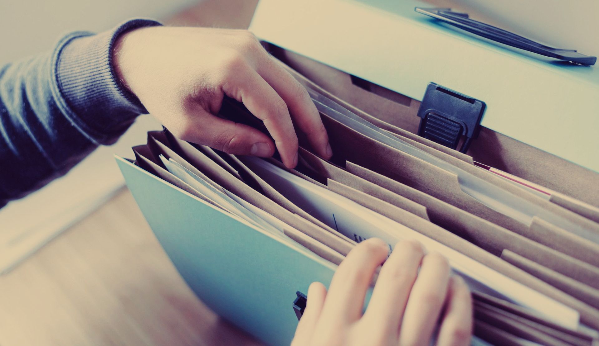 Hands sorting through files in a blue accordion file folder, with a utility knife visible in the background.