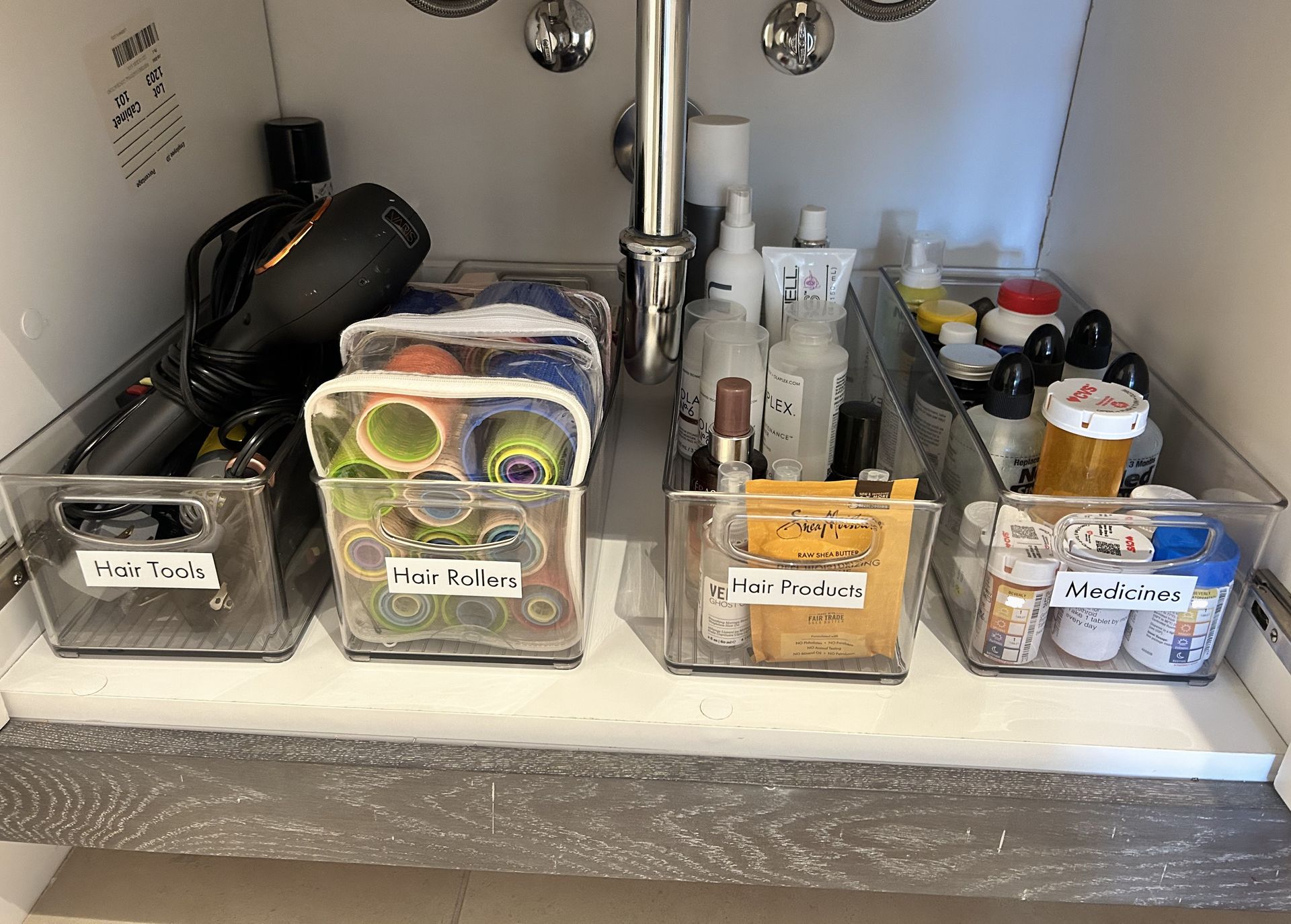 Bathroom cabinet with clear bins organizing hair dryer, hair products, toiletries, and medications.