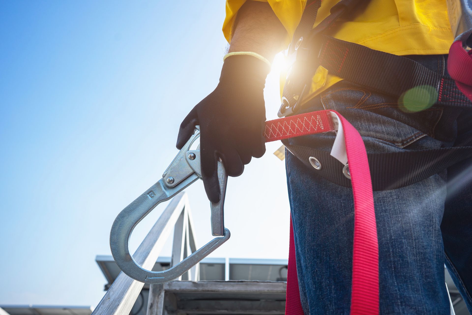 Worker in safety gear holding a carabiner with sunlight behind.