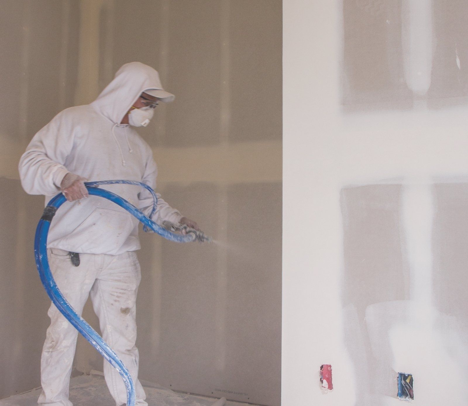 Man in protective gear sprays drywall mud indoors.
