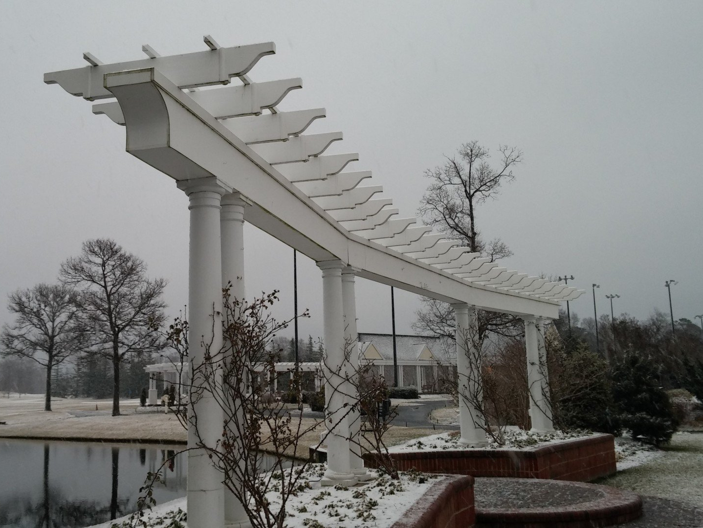 White pergola covered in snow, over a brick structure, near a pond with bare trees in background.