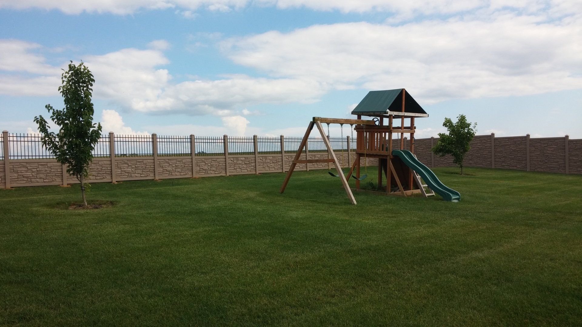 A backyard with a swing set, lawn, and fence under a partly cloudy sky.