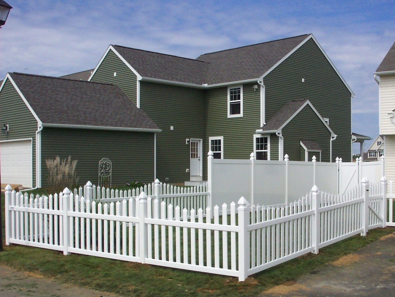 White picket fence around a green house with a dark gray roof, under a blue sky.