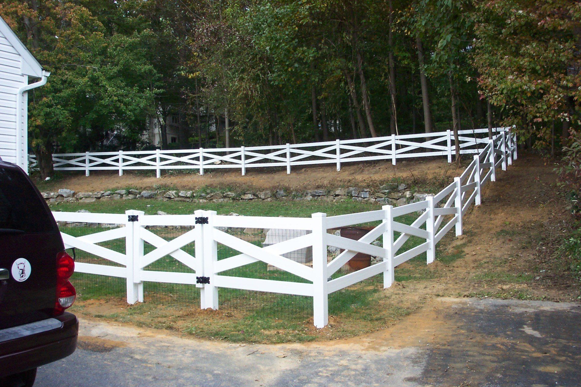 White picket fence around a grassy yard, next to a wooded area. A dark-colored vehicle is in the foreground.