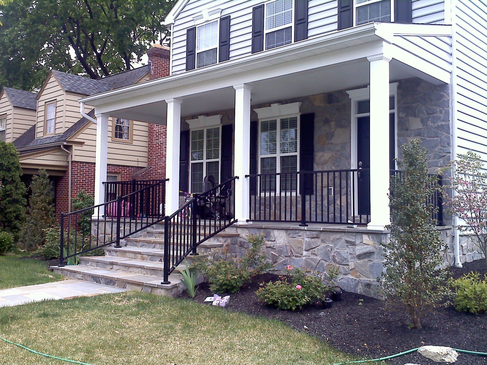 Front porch of a house with stone base, white pillars, black railing, and shutters.