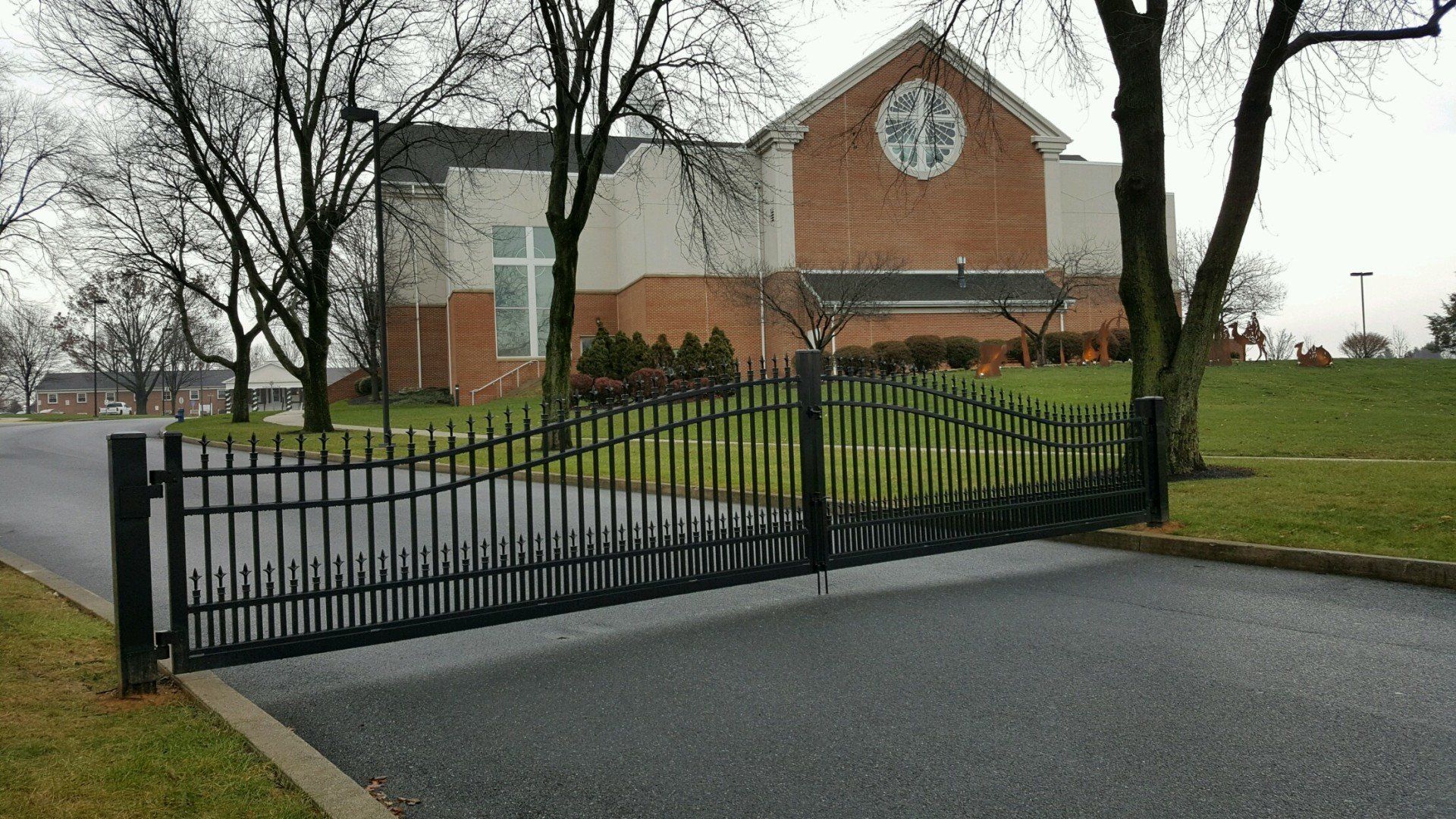 Black wrought iron gate on asphalt driveway, leading to a brick and white building with a round stained-glass window.