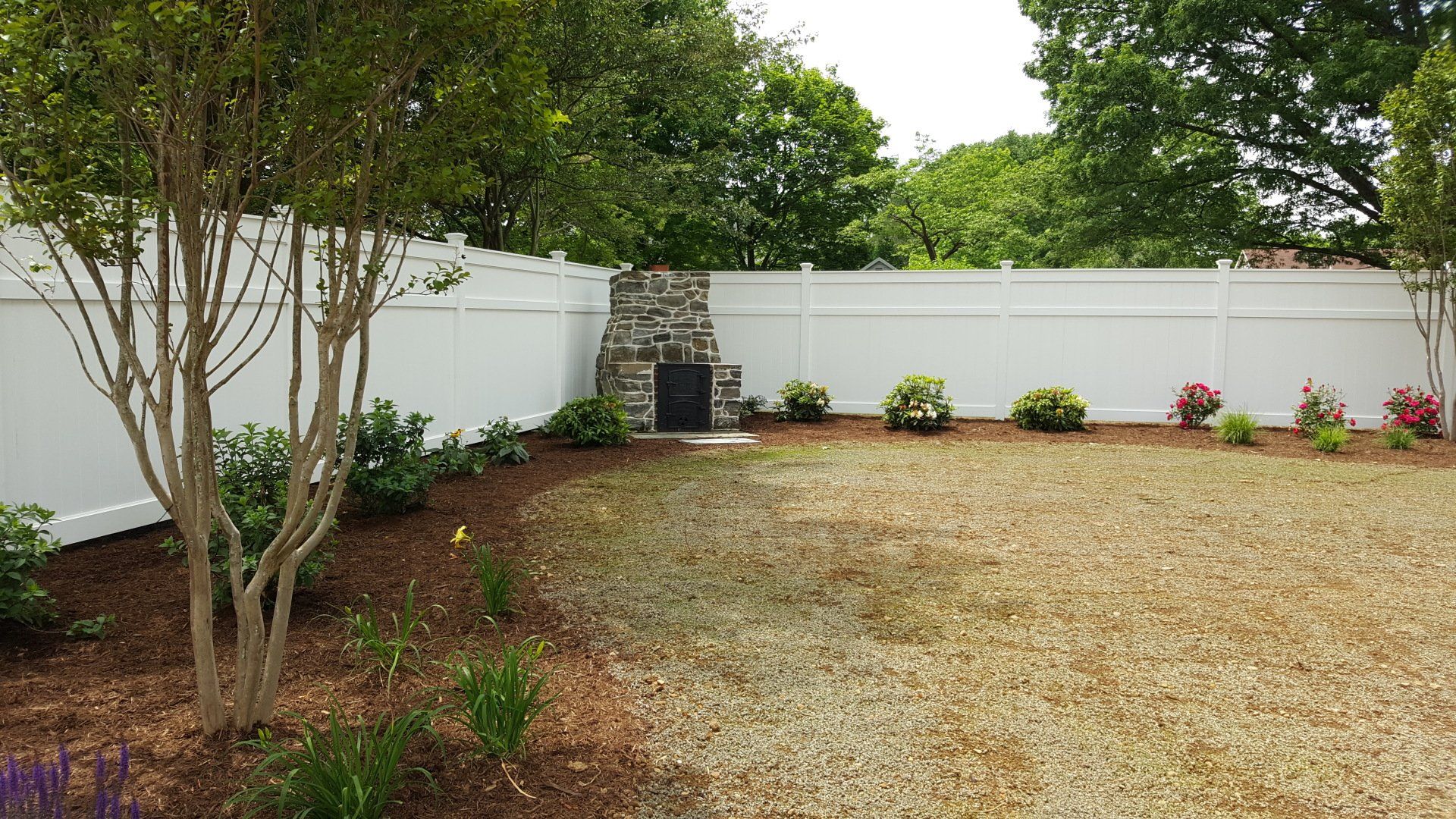 White fenced backyard with stone fireplace, landscaping, and brown mulch.