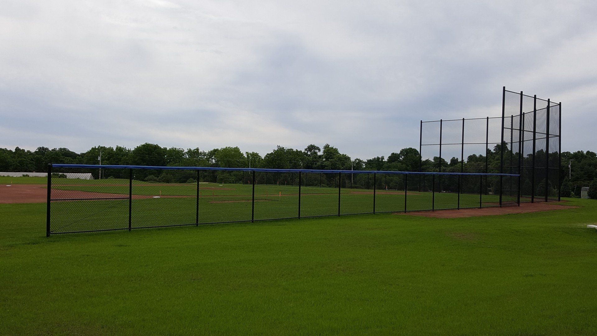 Baseball field with protective netting and green grass under an overcast sky.