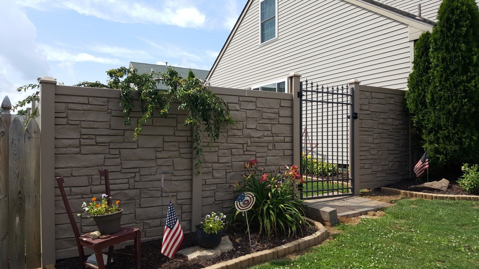 Stone-textured privacy fence with black metal gate and garden bed with American flags in front of a house.