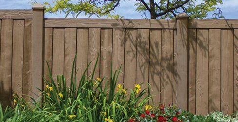 Brown wooden fence with tall green plants and flowers in front. Sunlight casts tree shadows.