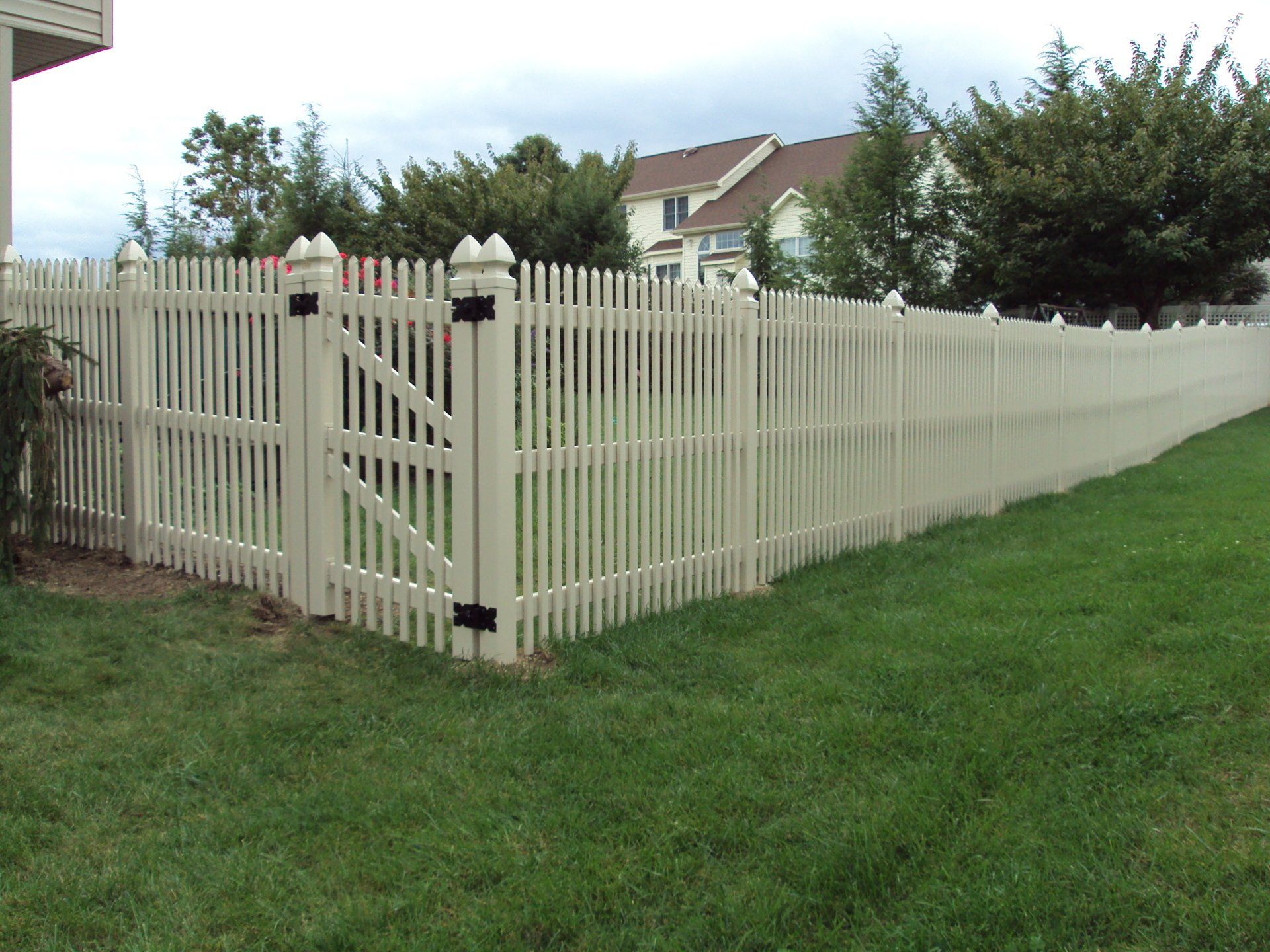 Beige picket fence enclosing a grassy yard, with a gate and a house visible in the background.